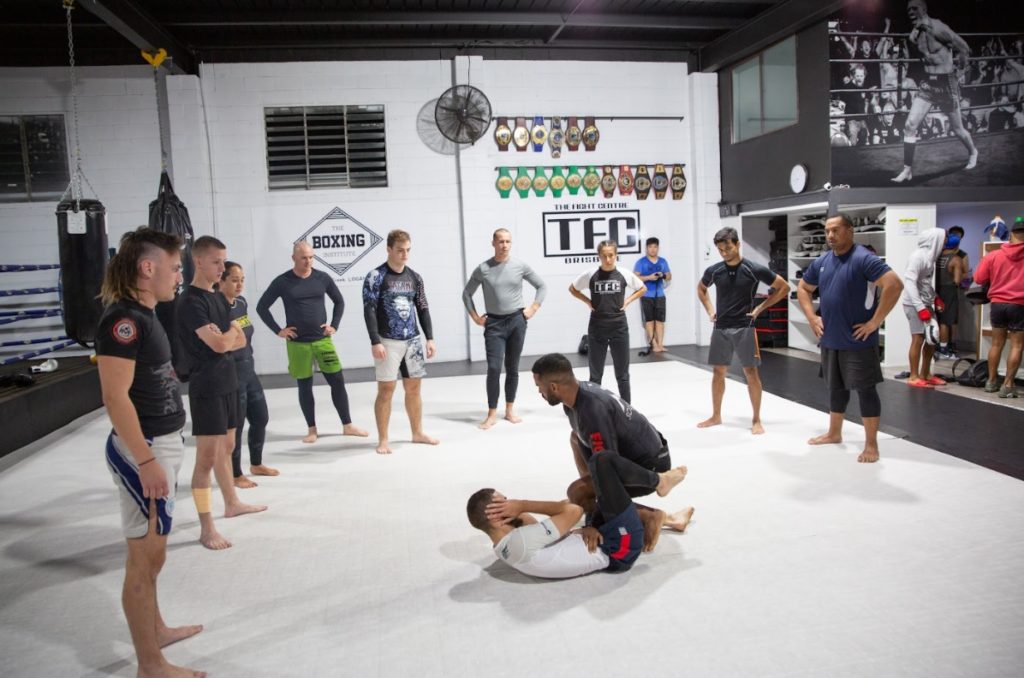 BJJ for beginners class. Two men demonstrate positions while other players stand in a circle to observe.