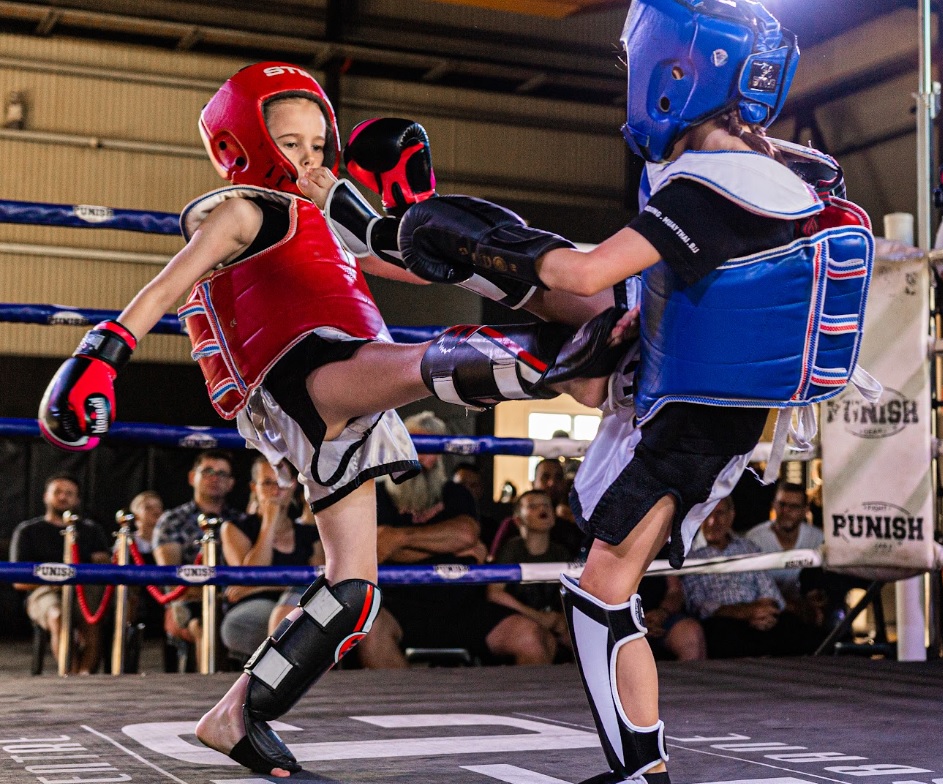Two young girls decked out in full safety gear (headgear, chest piece, and shin guards) engage in an exchange of strikes.